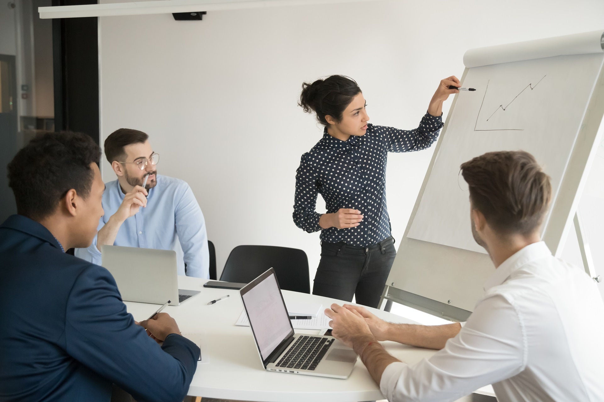 Multi-ethnic businesspeople gathered together in boardroom, staff listens lecture look at graph shown on flip chart, executive coach work with employees to help them achieve work-related goals concept